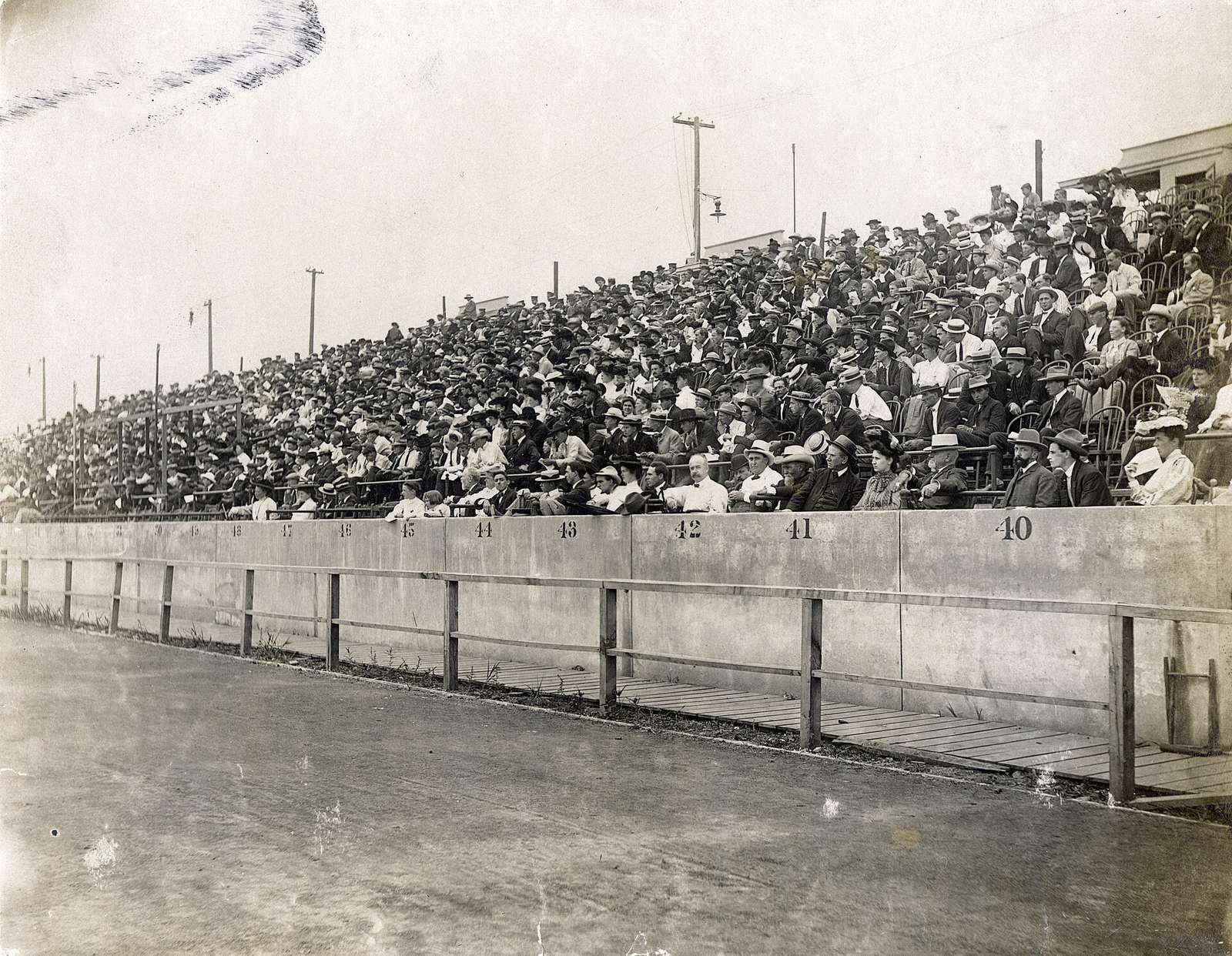 Black and white photograph of a packed grandstand at Francis Field in St. Louis during the 1904 Olympic Games, spectators in hats and white shirts above a numbered railing