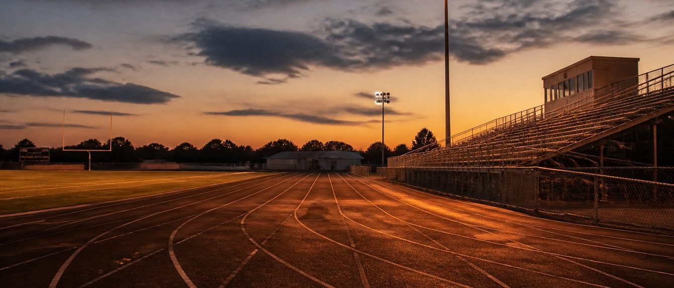 Empty stadium track at dusk, lane lines curving toward the far straight, one light tower lit above the bleachers